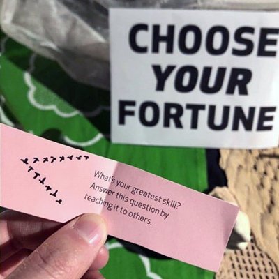 Close up of a hand holding a pink utopia fortune paper reading, 'What's your greatest skill? Answer this question by teaching it to others.'